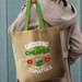 A jute tote bag with green handles, featuring a statement about gardening and tomatoes. It has a logo.