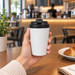 A reusable white coffee cup with a black lid, held in a hand, set against a café background.
