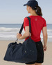 A woman wearing a red shirt and black shorts carries a navy blue duffle bag by the beach. The bag has a logo.