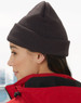 Acrylic beanie in dark grey worn by a person with long hair, paired with a red jacket. The beanie has a logo.