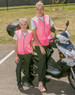 Two individuals wearing bright pink hi-vis safety vests stand next to a scooter. The setting appears to be outdoors.