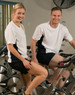 A white and navy short sleeve contrast tee worn by a man and woman sitting on stationary bikes in a gym.