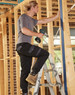 A pair of black work pants on a model using a power drill while standing on a ladder amidst wooden framing.