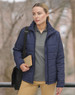 A woman in a navy puffer jacket stands outdoors, holding a book and wearing a grey polo shirt and tan trousers.