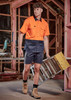 A man wearing an orange and navy work shirt and navy shorts carries a ladder in a construction setting. A man wearing an orange and navy work shirt and navy shorts carries a ladder in a construction setting.