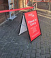 A-frame sign with a red background, featuring a logo and instructions for check-in entry. The frame is black.