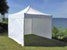 A white marquee tent stands on grass near a lake, featuring a peaked roof and open sides.