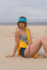 A model sits on the beach wearing a blue sports visor and yellow wrap, with a grey top and denim shorts.