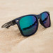 Black sunglasses with mirrored green and purple lenses, resting on sand. The sunglasses have a branding logo on the side. Black sunglasses with mirrored green and purple lenses, resting on sand. The sunglasses have a branding logo on the side.