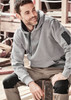 A grey unisex multi-pocket hoodie is worn by a man seated on a wooden bench, with worksite equipment in the background. A grey unisex multi-pocket hoodie is worn by a man seated on a wooden bench, with worksite equipment in the background.