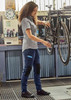 A woman in grey shirt and navy work pants is adjusting a bicycle in a workshop, featuring a logo on the pants. A woman in grey shirt and navy work pants is adjusting a bicycle in a workshop, featuring a logo on the pants.