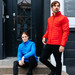 A red and a blue quilted jacket worn by a man and a woman, standing against a black door. The jackets feature a logo.