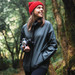 A woman wearing a dark green hooded rain jacket and a red beanie, holding a camera in a forest setting.