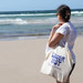 A person stands at the beach holding a natural-coloured tote bag with a gusset. The ocean waves are in the background. A person stands at the beach holding a natural-coloured tote bag with a gusset. The ocean waves are in the background.