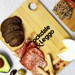 A large bamboo cutting board with assorted food items, including bread, meats, cheese, and an avocado, featuring a logo. A large bamboo cutting board with assorted food items, including bread, meats, cheese, and an avocado, featuring a logo.