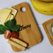 A small bamboo cutting board with cheese, tomatoes, mint leaves, and snacks, featuring a logo, beside ripe bananas. A small bamboo cutting board with cheese, tomatoes, mint leaves, and snacks, featuring a logo, beside ripe bananas.