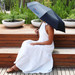 A black umbrella with a logo, held by a person in a white dress, sitting on wooden steps with greenery in the background. A black umbrella with a logo, held by a person in a white dress, sitting on wooden steps with greenery in the background.