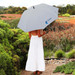 A grey umbrella with a logo is held by a person in a white dress, surrounded by garden scenery. A grey umbrella with a logo is held by a person in a white dress, surrounded by garden scenery.