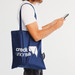 A navy blue tote bag with a logo sits over the shoulder of a person holding a smartphone. A navy blue tote bag with a logo sits over the shoulder of a person holding a smartphone.