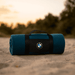 A rolled teal blanket with a black strap and logo, placed on sandy ground against a sunset backdrop.