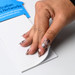 A hand with long, patterned nails peels back a sheet from a white, lined rectangular notepad with a blue branding element.