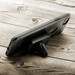 A black phone holder with a triangular base, positioned on a wooden surface. It has a branding logo.