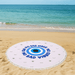 A round beach towel in white and blue with a central eye design, placed on a sandy beach by the water.