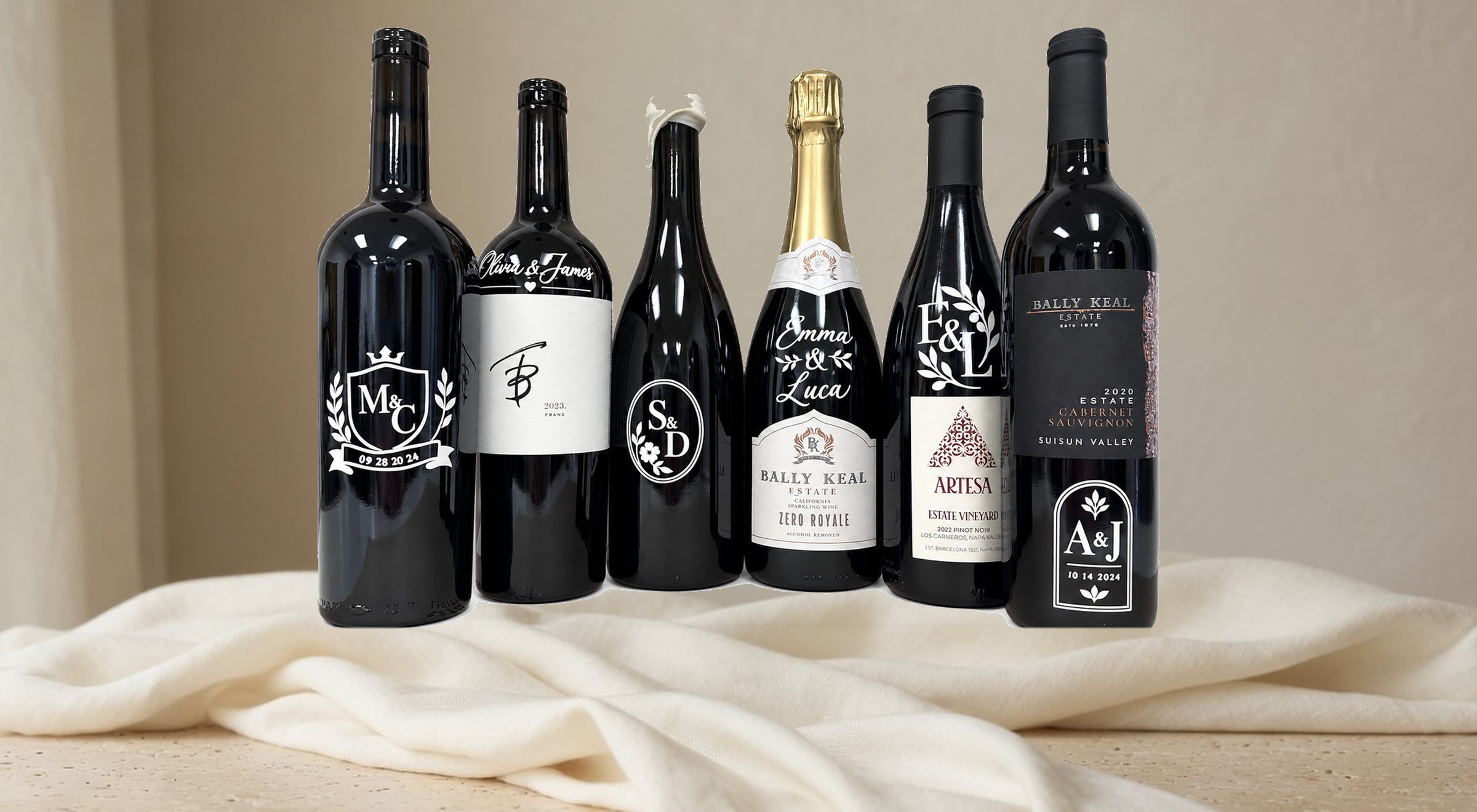 Six personalized bottles standing on a light linen cloth: M and C, Olivia and James, S and D, Emma and Luca, E and L on Artesa, and A and J on Bally Keal Cabernet.
