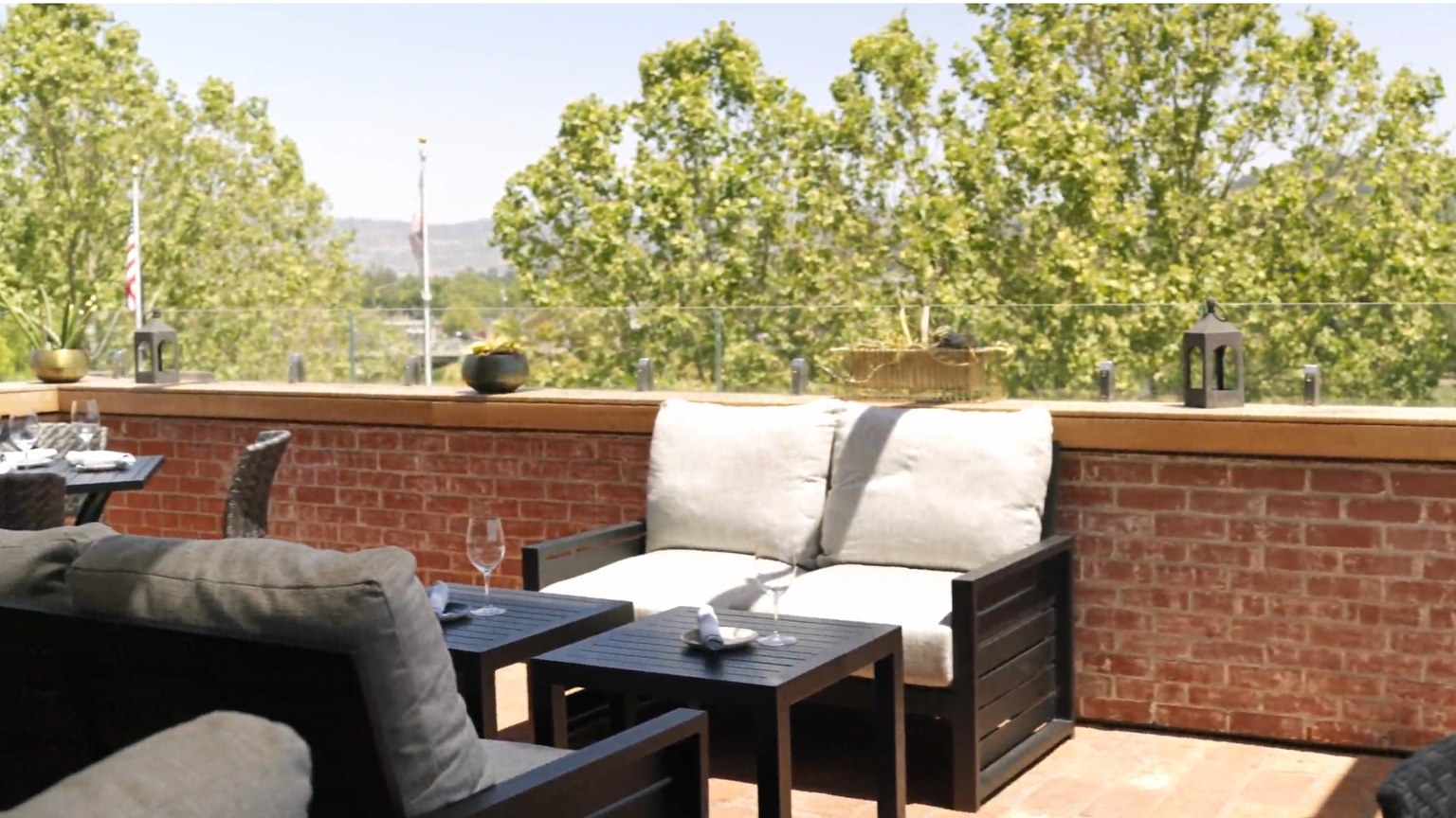 A brick terrace with outdoor sofas, wine glasses, and trees beyond the rail.