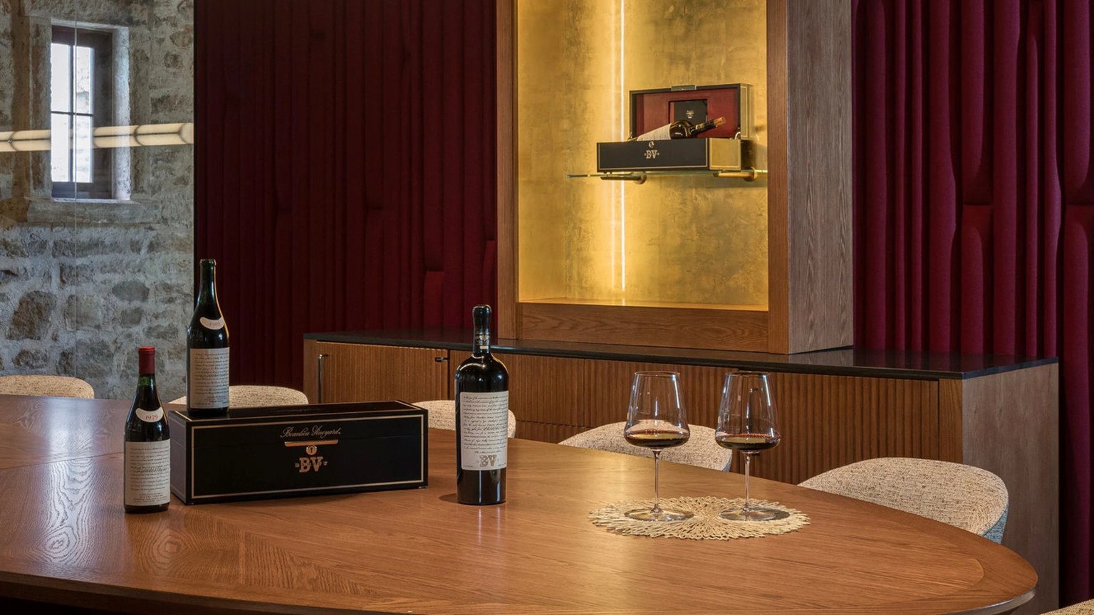 An oval oak table set with Georges de Latour bottles and glasses against a crimson wall