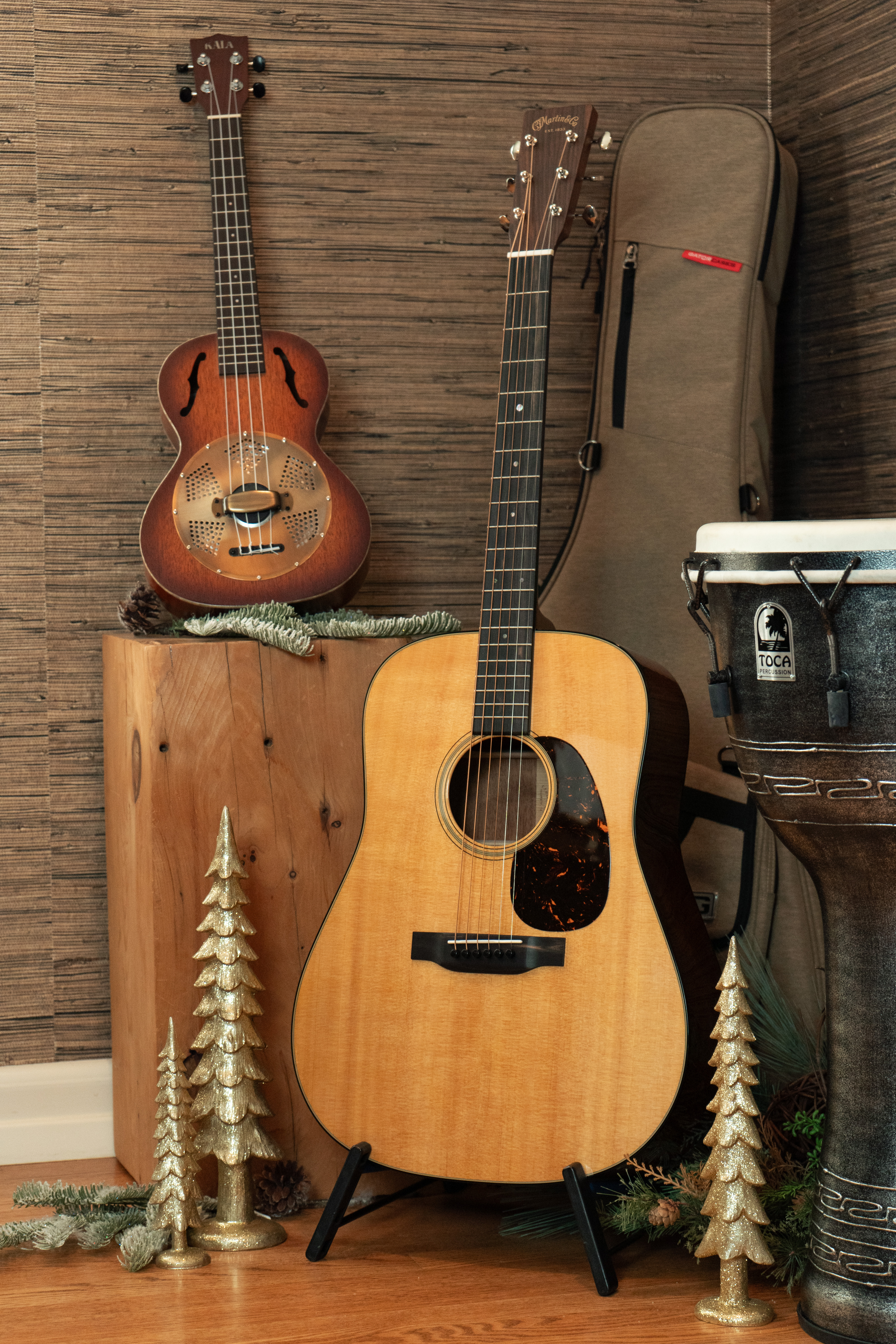 A Martin D-18 acoustic guitar on a stand with a Kala ukulele and Toca djembe surrounded by subtle holiday decorations.