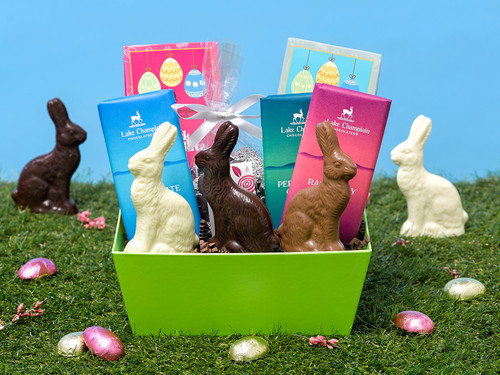 Assorted Easter chocolates packaged in a lime green reusable cardboard basket, sitting on a background of grass and a blue sky with pink flowers, wrapped chocolate eggs and chocolate bunnies in the background.