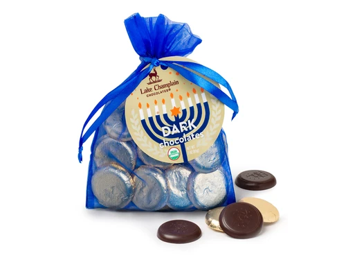 Organic dark chocolate Hanukkah coins wrapped in silver foil and packaged in a royal blue organza gift bag with wrapped and unwrapped chocolate coins around the bottom of the right side of the bag.