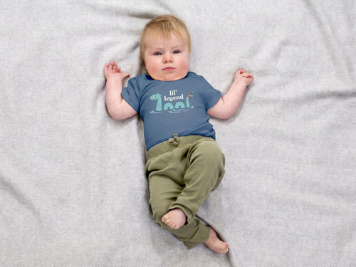 Baby boy laying on a light gray blanket wearing sage green pants and a Pacific blue 12-18 month old size organic cotton onesie with the lake monster Champ on the front holding a piece of chocolate at the end of his tail and the words "Lil Champ" above Champ.