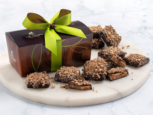 Dark Chocolate maple pecan butter crunch pieces on a ceramic white plate with a brown gift box and a green and brown ribbon, in the background.