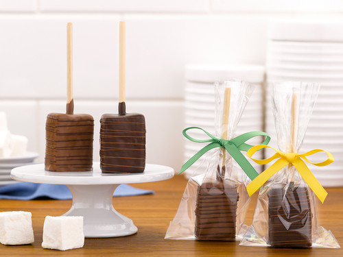 Assorted milk and dark chocolate covered marshmallow pops on a wooden table with two unwrapped on a white cake stand and two next to the stand individually wrapped in a clear gift bag, milk chocolate with a green ribbon and dark chocolate with a yellow ribbon, with 2 marshmallow squares on the left in front of the cake stand and part of a square marshmallow showing in the background.