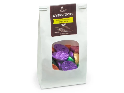 White paper bag with a clear window showing chocolate eggs wrapped in assorted colorful foils and a brown label that says "overstock eggs" on it.
