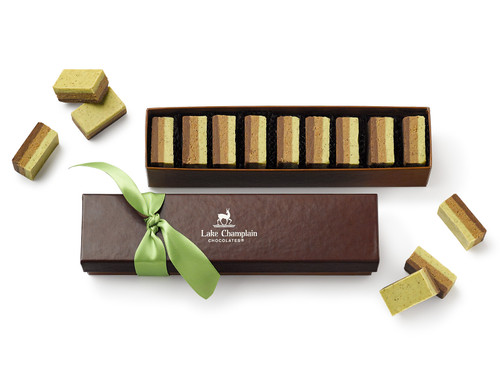 Pieces of pistachio praline laying in a brown gift box with the lid open and in front of the gift box, with a green ribbon on the lid and pieces of pistachio praline around the box View Product Image