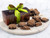 Dark Chocolate maple pecan butter crunch pieces on a ceramic white plate with a brown gift box and a green and brown ribbon, in the background. View Product Image