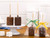 Assorted milk and dark chocolate covered marshmallow pops on a wooden table with two unwrapped on a white cake stand and two next to the stand individually wrapped in a clear gift bag, milk chocolate with a green ribbon and dark chocolate with a yellow ribbon, with 2 marshmallow squares on the left in front of the cake stand and part of a square marshmallow showing in the background. View Product Image