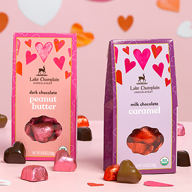 Dark peanut butter hearts gift box and milk chocolate caramel great gift box sitting on a pick table with wrapped and unwrapped hears around the boxes and paper hearts in the background.