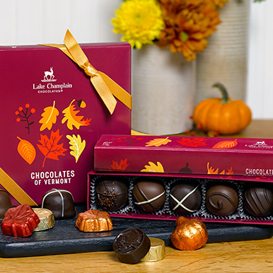Autumn Chocolates of vermont gift box standing up next to the Autumn 5 piece truffle gift box with the lid sitting on top of the truffle gift box and assorted autumn colored flowers in a white vase  and a small pumpkin next to it in the background and assorted wrapped and unwrapped chocolate leaves and pumpkins sitting in front of the gift boxes.