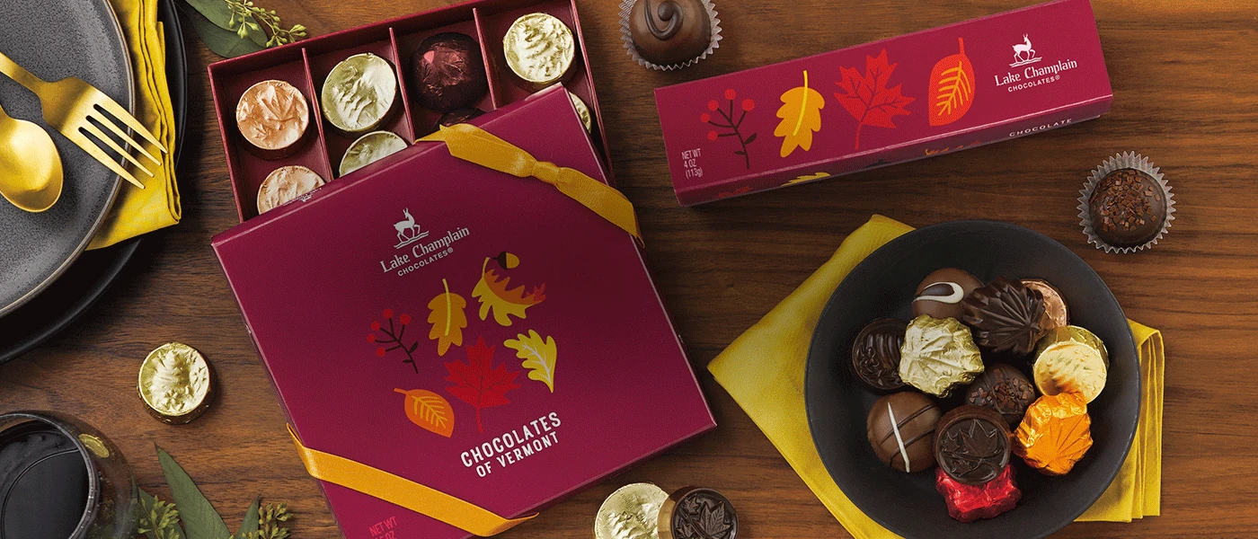 Assorted fall themed chocolate gift boxes on a wooden table  with wrapped and unwrapped chocolates in and around the boxes, and a round dark gray plate with assorted fall chocolates sitting on a golden color napkin and a gray plate with gold utensils in the left hand corner.