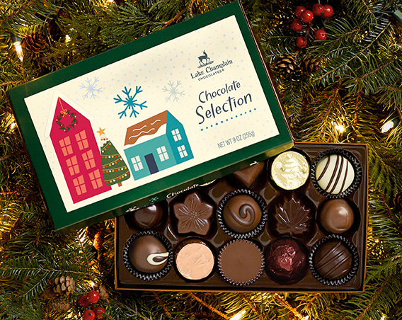 Assorted gourmet chocolates in a holiday themed gift box sitting on greenery with berries and white Christmas lights.