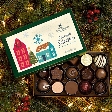 Assorted best selling chocolates in a holiday themed gift box, sitting on Christmas greenery with holiday lights, pine cones and berries.