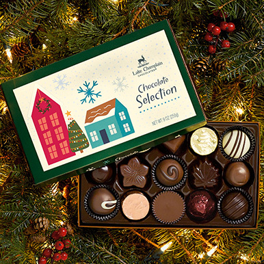 Gourmet chocolates in a holiday gift box sitting Christmas tree limbs with white Christmas lights.