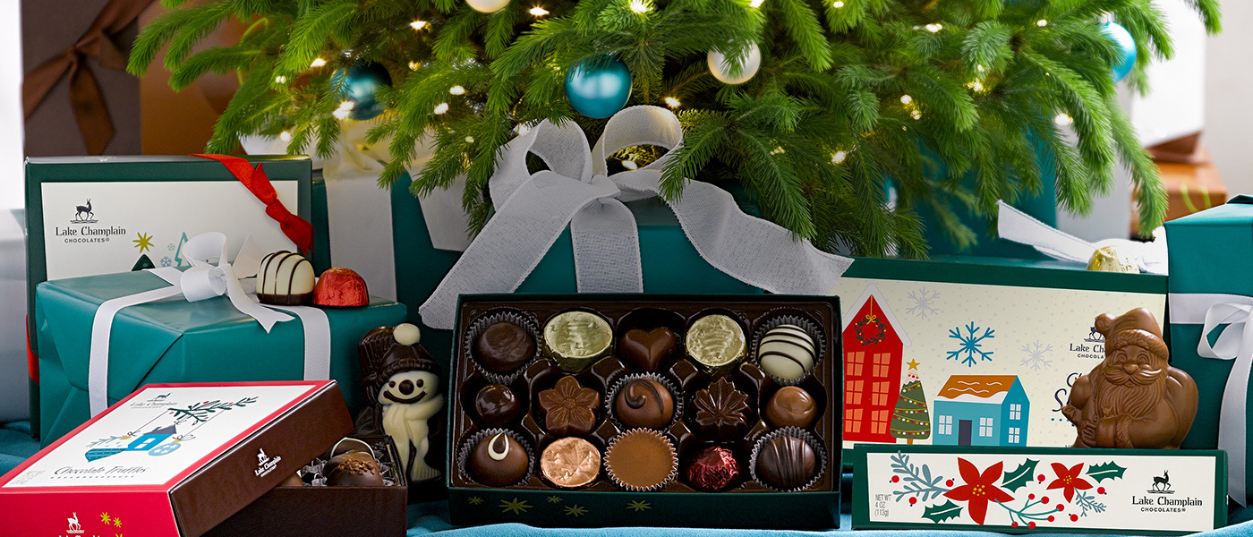 Assorted Christmas chocolates and Christmas gift boxes under a Christmas tree with blue and cream balls, a cream bow, and white Christmas lights.