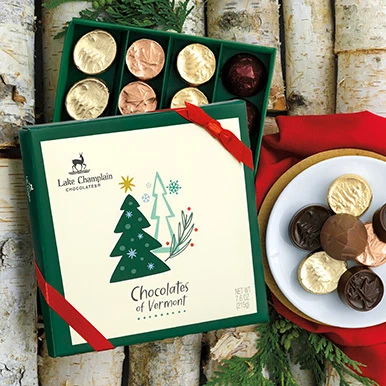Assorted Chocolates of Vermont in a holiday themed gift box with red ribbon, sitting on pieces of birch trees with a round white dish full of wrapped and unwrapped chocolates sitting on top of a gold dish and a red napkin, with cedar greenery around the top right of the gift box and under the plate of chocolates.