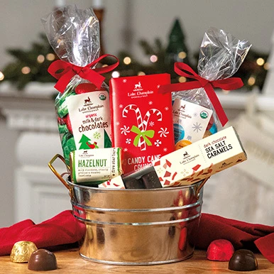 Assorted Christmas chocolates in a holiday gift tin sitting on a wooden table with a red table cloth behind the tin with wrapped and unwrapped chocolates next to the gift basket.