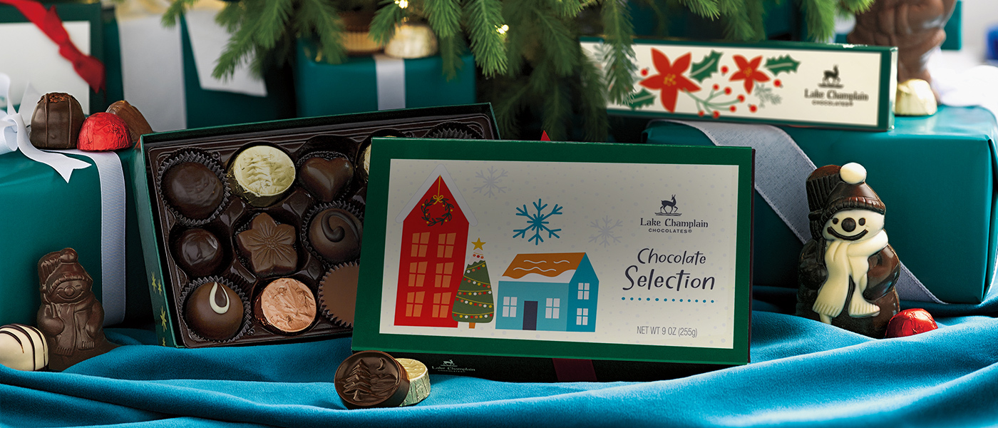 Assorted Christmas chocolates in Christmas themed gift boxes with a dark chocolate snowman under a Christmas tree with teal-blue and cream decorations on it and teal and white packages under it.